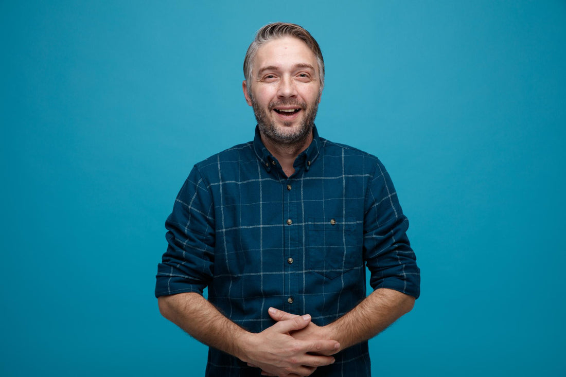 Middle age man with grey hair in dark color shirt laughing out holding hands on his belly happy and joyful standing over blue background